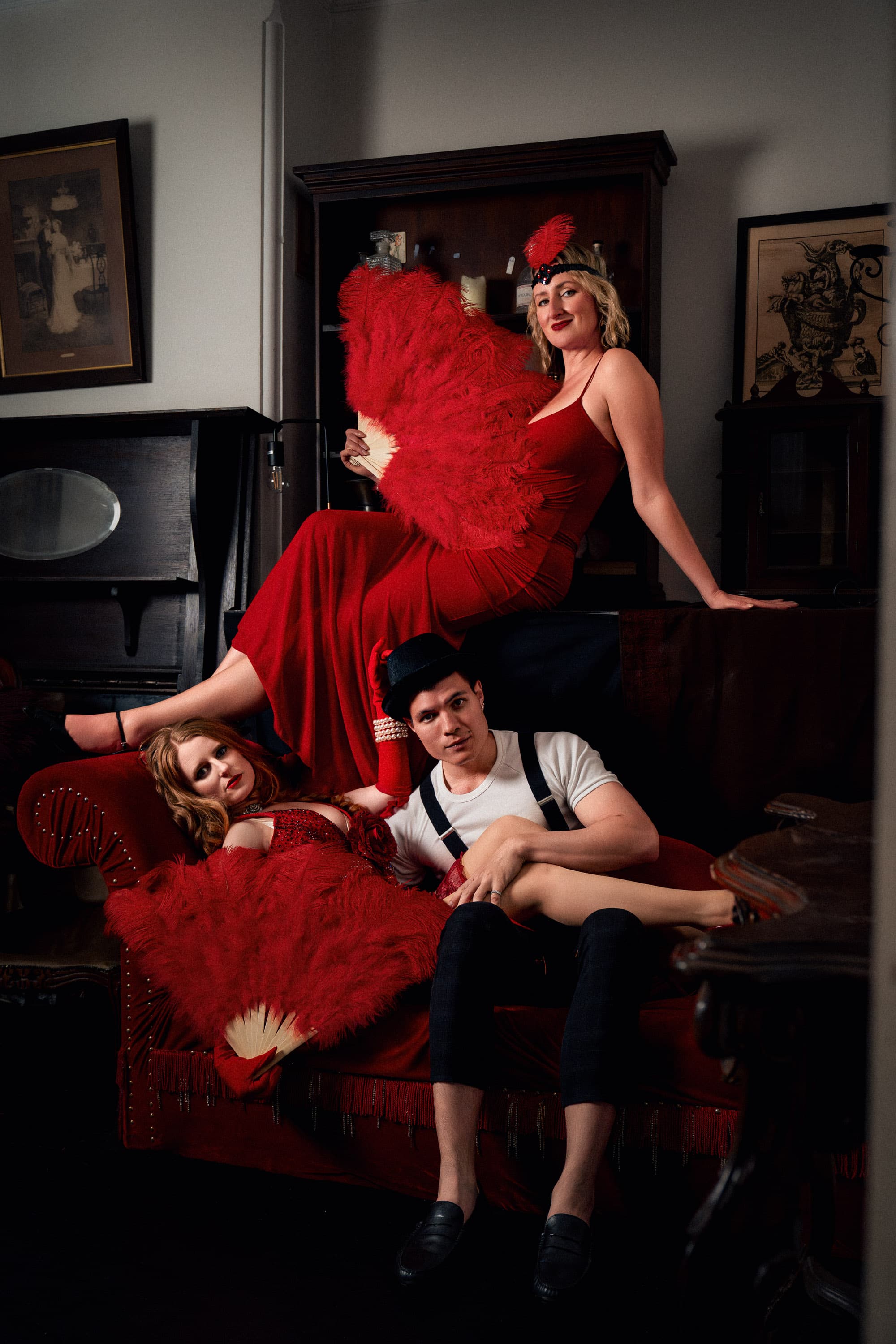 Three cabaret performers on a red sofa. Two women wearing a red dress, one man wearing a white tshirt.
